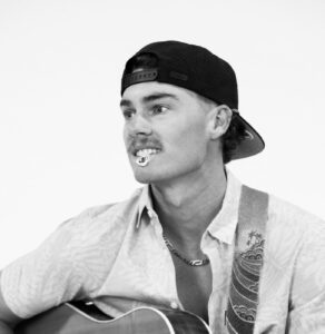 Black and white photo of carter smith holding a guitar, wearing a white shirt and a backwards black hat