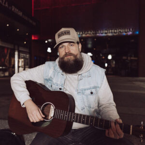Matt burke wearing a tan hat that says nash, a jean vest and gray hoodie, holding a brown acoustic guitar