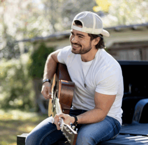 Emmet Stevens, Jr. wearing a white t-shirt and white hat holding a guitar on the back of a truck bed